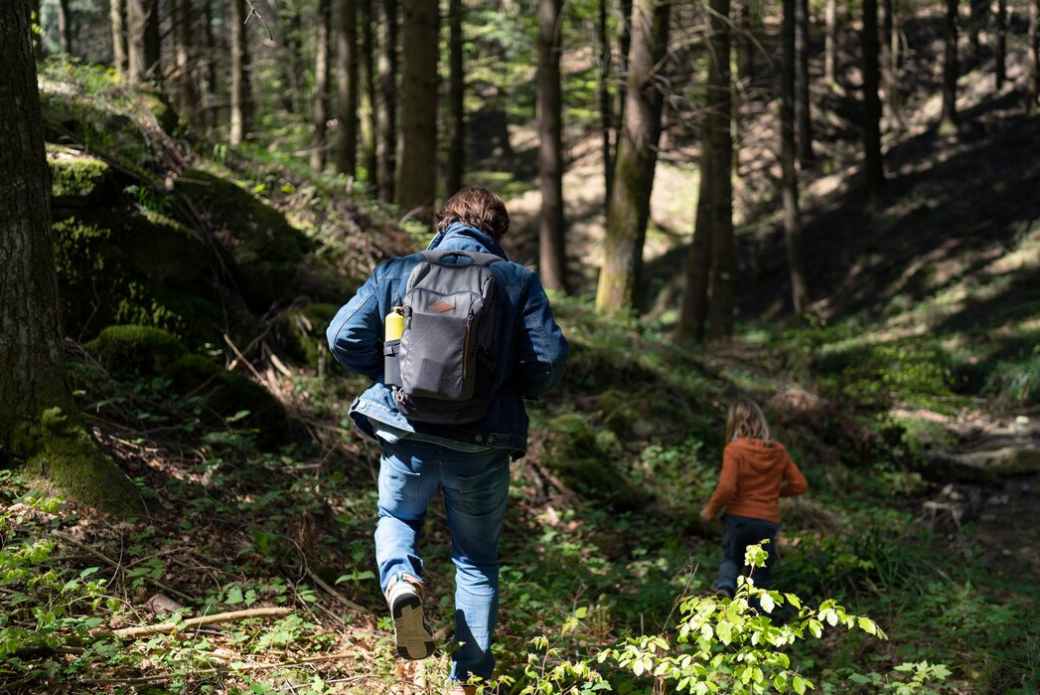 A person with a backpack hikes through a forest, followed by a child.