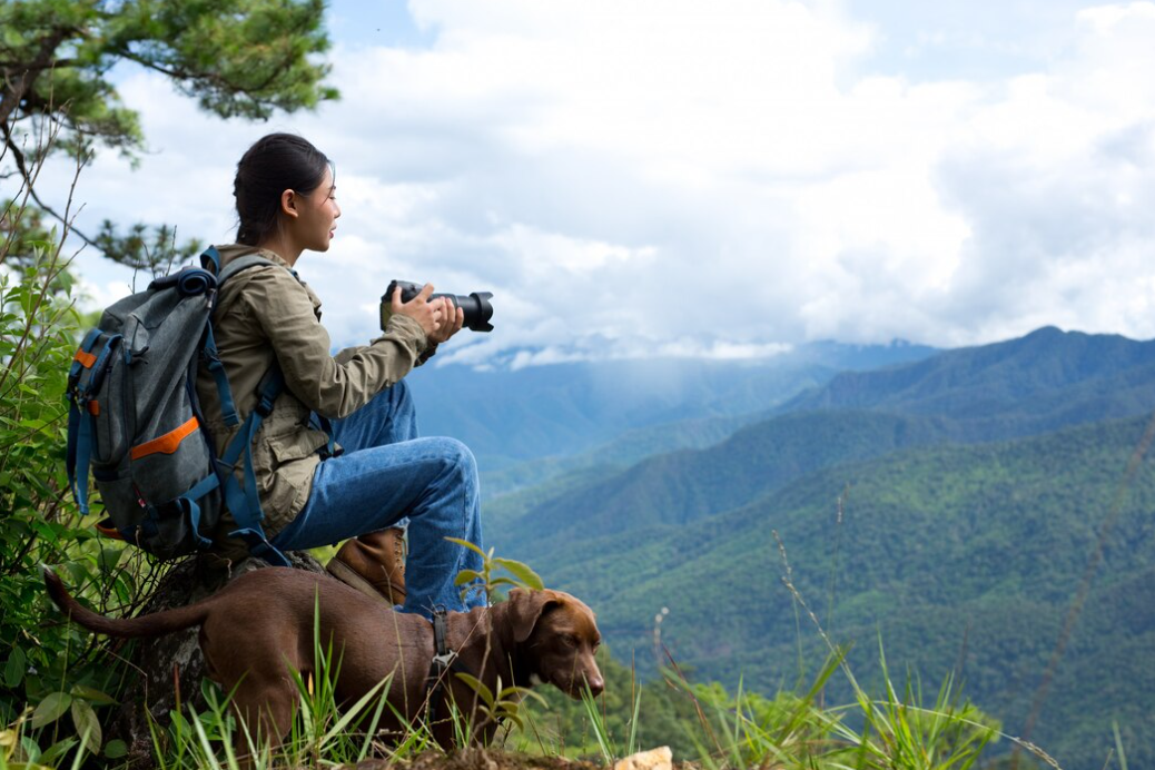 A woman with a backpack sits on a mountain's edge, looking out with her camera, dog stands nearby.