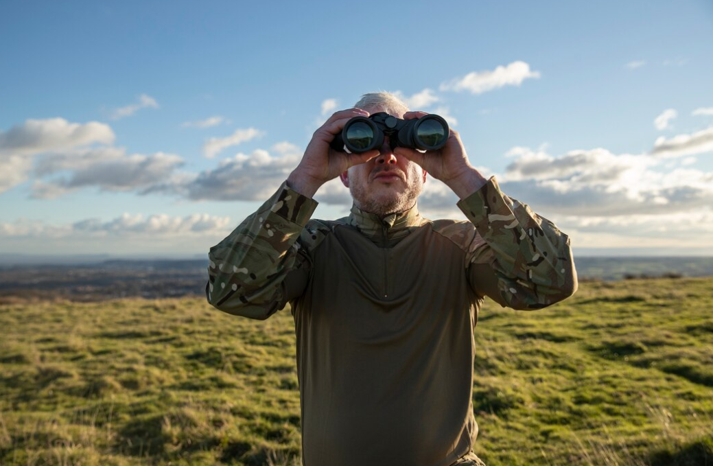 man in military clothing looks into binoculars and holds it with two hands, the filed behind him